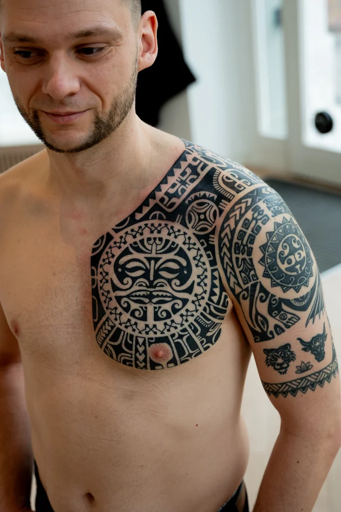 Marquesas-inspiriertes Tattoo auf Brust, Schulter und Oberarm mit figurativen Rundmotiven und Musterbändern